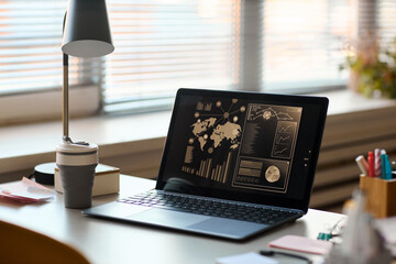 Open laptop displaying data analytics dashboard with world map and charts on screen, sitting on office desk near reusable coffee cup, lamp, and stationery, blinds covering window