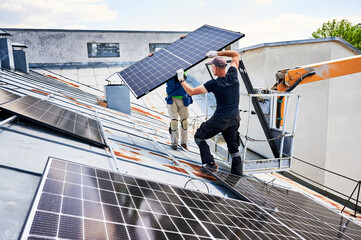 Workers building solar panel system on metal rooftop of house with assistance of crane lift. Two men installers carrying photovoltaic solar module outdoors. Renewable energy generation concept.