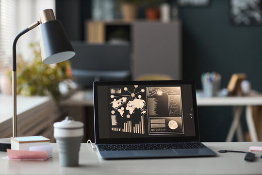 Modern workspace featuring open laptop displaying business analytics dashboard with world map, charts, and graphs on screen, desk lamp and office supplies surrounding device - Powered by Adobe
