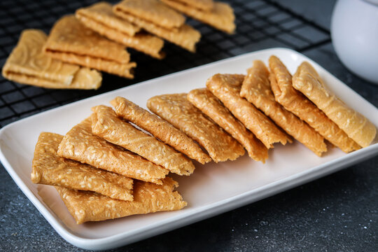 A view of crispy, golden-brown Kue Semprong or Love Letter cookies, a thin, folded Southeast Asian snack. Arranged on a white plate and a cooling rack, highlighting their delicate texture.