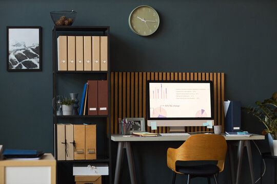Modern office workspace featuring computer monitor displaying business charts and graphs on desk with organized folders and binders on shelves against dark wall background