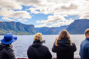 Tourists stand on a boat deck viewing the massive cliffs of Western Brook Pond fjord in Newfoundland, Canada, under a dramatic cloudy sky during the autumn season.