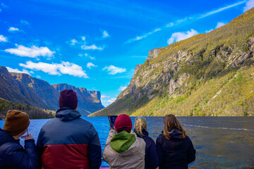 Passengers on a boat tour photograph the steep, sunlit rock walls of Western Brook Pond in Gros Morne National Park, showcasing vibrant fall foliage and blue skies.