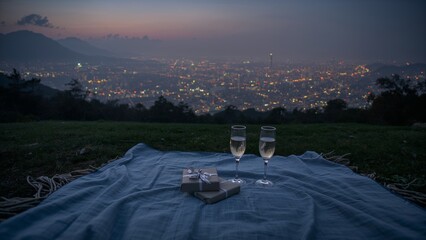Romantic twilight picnic: Champagne, gifts, and sparkling city lights from a hilltop view.