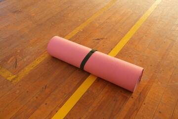 A pink yoga mat is laying on a wooden floor. The mat is unrolled and has a green strap tied to it. Concept of relaxation and tranquility, as the yoga mat is often associated with yoga