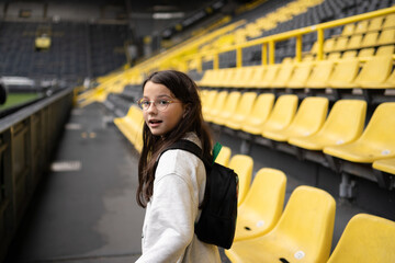 Smiling teenage girl in glasses with backpack on empty football stadium, education travel and leisure concept