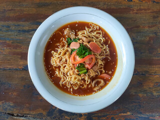 Bowl of noodles in savory soup with sausage slices, tomato, herbs, and sesame seeds on rustic table.