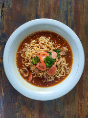 Bowl of noodles in savory soup with sausage slices, tomato, herbs, and sesame seeds on rustic table.