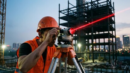 Young construction worker wearing a hard hat and safety vest operating a laser level with a bright red beam on a contemporary high-rise building construction site during evening - Powered by Adobe