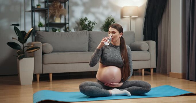 Pregnant woman in yoga attire sits cross-legged on mat, drinking water, holding belly, indoors with sofa and plant. Pregnant Woman Drinking Water Yoga