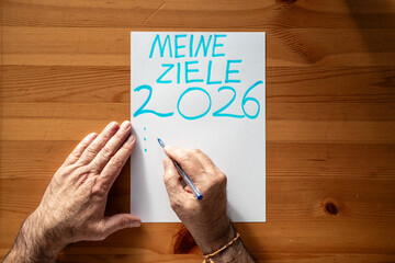 Meine Ziele 2026 H&auml;nde schreiben Neujahrsvors&auml;tze auf Papier, leere Liste f&uuml;r pers&ouml;nliche Planung, Motivation und Neubeginn auf Holztisch