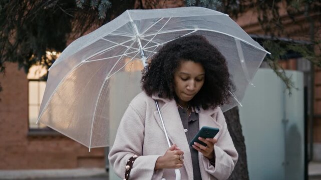 Young woman checking social network while standing with transparent umbrella, waiting for her boyfriend