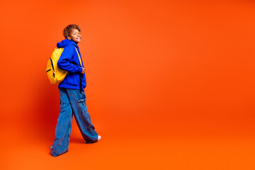 Energetic young boy smiles while wearing blue hoodie and yellow backpack walking against a bright orange background