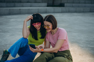 Joyful young women with quad skates watch videos via smartphone on ground

