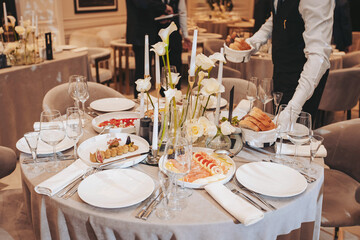 A table is set with multiple plates of food, glassware, and flower arrangements. A server is placing more food while guests socialize in the background at a restaurant during an event