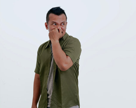 Asian man isolated over white background smelling something stinky and disgusting, intolerable smell, holding breath with fingers on nose. Bad smells concept.