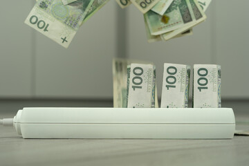Polish government is raising electricity prices in Poland. Power supply strip (cord) with 100 zloty banknotes symbolizes rising prices. Flying PLN banknotes and office environment as background