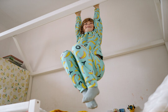 Child hangs from a loft bed in a bedroom wearing banana patterned pajamas