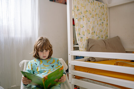Boy sits in bedroom reading a book while wearing pajamas