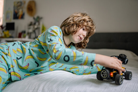 Boy plays with toy car in bedroom while wearing pajamas and lying on bed