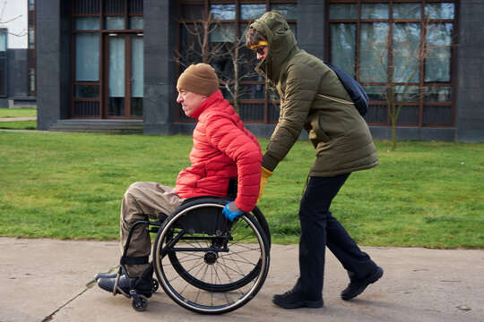 A man in a wheelchair is riding on the sidewalk while a woman is helping him.