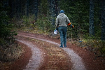 Obraz premium Male treasure hunter using metal detector on winding forest path, surrounded by tall trees and lush greenery, exploring nature for hidden treasures and artifacts