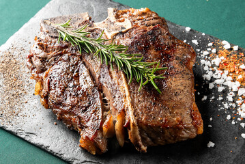 A perfectly grilled T-bone steak on a black slate board, seasoned with salt, pepper, and fresh rosemary sprigs for a delicious meal