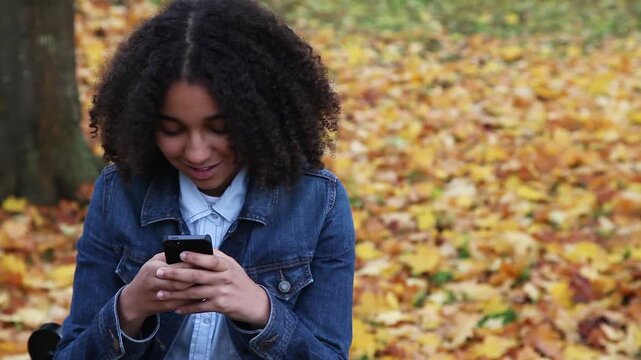 Beautiful mixed race African American girl teenager young teen wearing a blue denim jacket outside happy and smiling using her cell phone for social media
