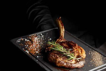 A tomahawk steak with rosemary and spices on a black serving board. This close-up, dramatic shot in...