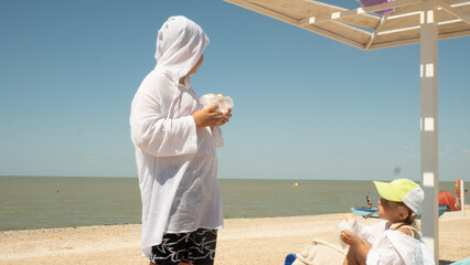 Two children in white shirts eat snacks on a sunny sandy beach under an umbrella, admiring the calm ocean during a family summer vacation. The children snack on the beach between swims.
