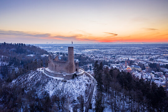 Aerial view of Windeck Castle ruins atop a snow-dusted hill, silhouetted against a vibrant sunset sky, with the town nestled below., Weinheim, Baden-Wurttemberg, Germany.