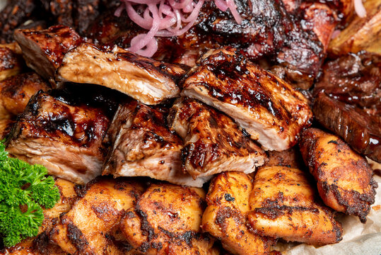 A close-up shot of a hearty mixed barbecue platter featuring glazed pork ribs, chicken pieces, and sausage links, garnished with pickled red onions and fresh parsley