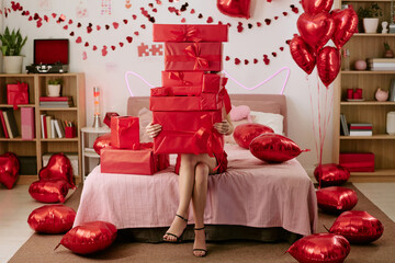 Woman sitting on bed holding large stack of red gift boxes covering face surrounded by heart shaped balloons and Valentines Day decorations in bedroom