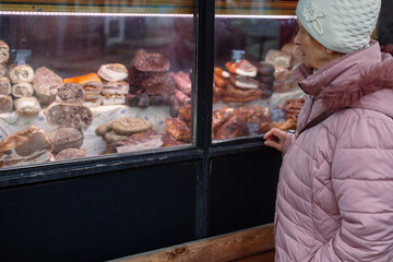 Senior woman in winter clothing inspecting glass display of cured meats and sausages at butcher shop. Everyday shopping and meat consumption.