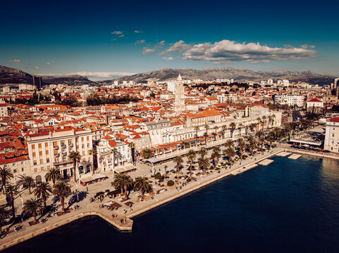 Aerial view of terracotta rooftops meet the azure Adriatic Sea, with palm trees lining the Riva promenade against the backdrop of distant mountains, Split, Split-Dalmatia County, Croatia.