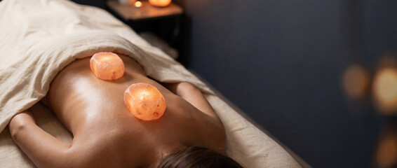 Relaxing spa massage with warm glowing Himalayan salt stones on a woman's back. Wellness therapy concept in a dark atmosphere with copy space for text.