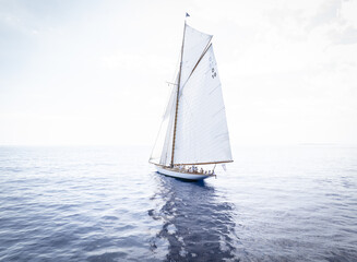 Palma, Spain - 16 August 2024: Aerial view of a sailing boat cutting through the deep blue sea, its white sails billowing against the bright sky, a dance of light and shadow on the water's surface.