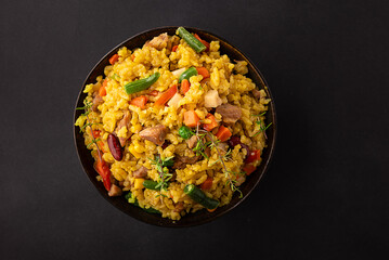 Stewed rice with meat and assortment on vegetables and spices in dark ceramic bowl on black stone table top view. Tasty homemade food concept.