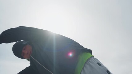 low-angle silhouette caucasian goalkeeper with arms raised against bright sunflare, dramatic backlight and cold sky, hoodie and beanie rimmed by light, triumphant stance on empty urban pitch
