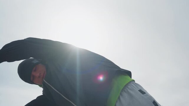 low-angle silhouette caucasian goalkeeper with arms raised against bright sunflare, dramatic backlight and cold sky, hoodie and beanie rimmed by light, triumphant stance on empty urban pitch