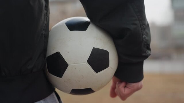tight frame of soccer ball tucked against hip, white player stands beside goal with relaxed posture on community pitch, contemplative mood suggests halftime pause or plan before restart