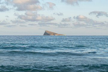 Benidorm Island seen from the sandy beach, with a large cruise ship offshore, small boats and a sailboat on the Mediterranean Sea under a blue sky dotted with white clouds.