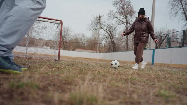 white grandmother practicing penalty near small goal in cold urban park, brown coat and beanie, focused kick across patchy grass, casual shoes and worn net, low camera angle captures motion