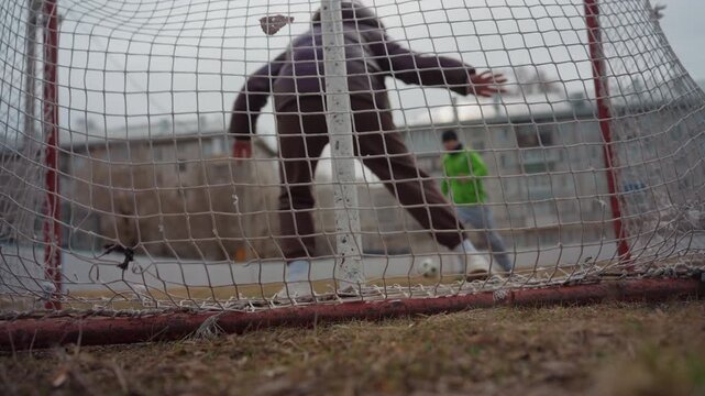 low net view of save attempt, silhouette of adult leaping and child striker in green approaching; ball meets mesh against overcast sky, gritty urban playground tension and dynamic motion for sports