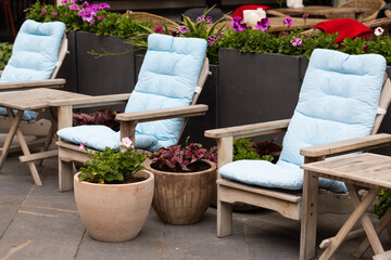 Chairs with blue cushions are placed next to tables and flower pots. Green plants surround the area, showing a lively outdoor setting on a sunny day.