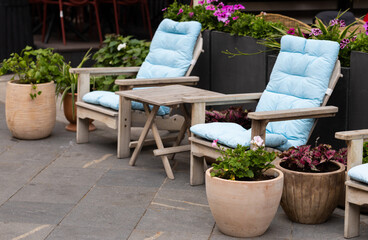 Two relaxed chairs with light cushions sit next to a small table. Flower pots surround them on a paved patio during a bright afternoon in a garden space.