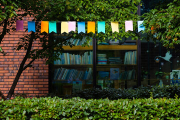 Colorful flags are strung above a library surrounded by trees and shrubs. The library has glass windows with shelves of books visible inside.