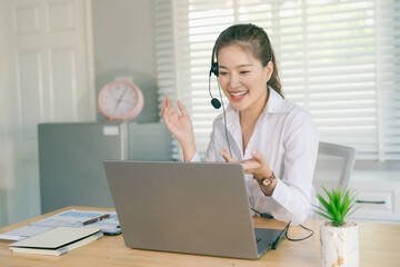 An Asian woman, a call center employee, wears headphones, works from home, communicates via...