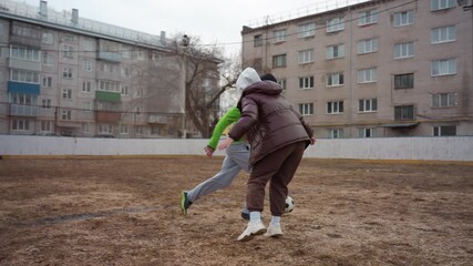 friendly soccer duel between white man and white senior woman on muddy courtyard pitch, passing and tackling exchanges, apartment block backdrop, casual jackets and sneakers, playful competition