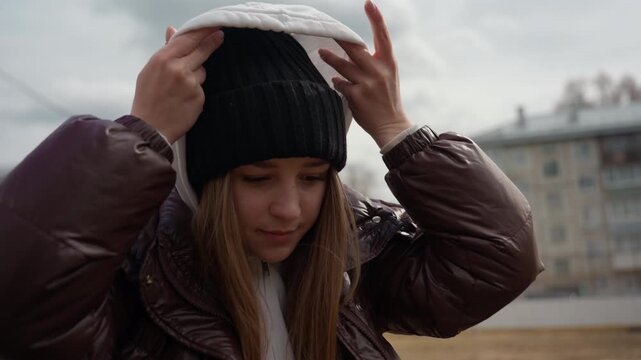white woman straightening black beanie and pulling hood up, brown coat and layered hood visible, subtle prep for chilly overcast walk, candid suburban street backdrop, focused expression.